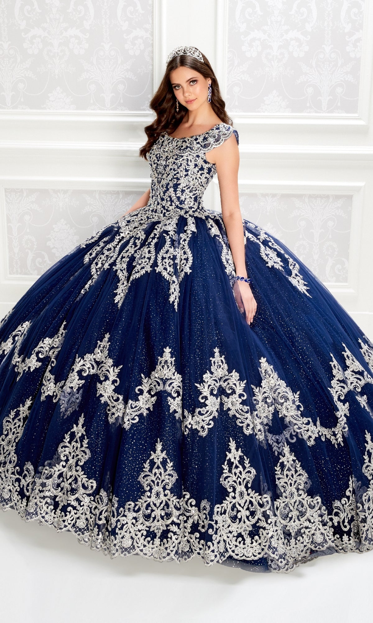A woman in the Quinceanera Dress PR22035 By Princesa, a navy blue gown with silver embroidery and a corset bodice, stands before a white ornate wall, holding out her skirt.